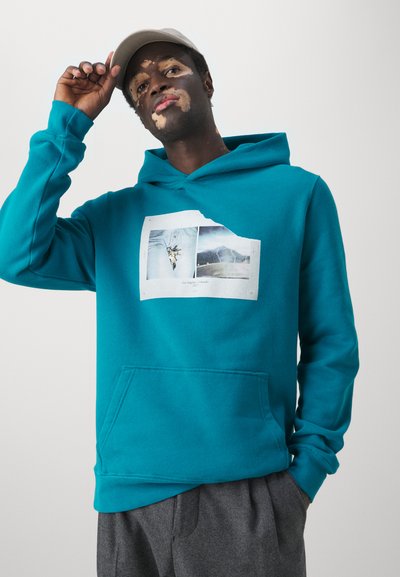 Teal hoodie made of soft cotton, featuring a front pocket and a graphic print with landscape images. The model wears a light gray cap.