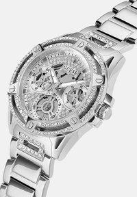 Srebrna ura iz nerjavečega jekla z okrasnim okvirjem, zapletenim številčnico in tremi podštevilčnicami. Ima polirano zapestnico z vrstico kristalov.