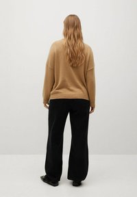 Beige neulottu villapaita, jossa on roikkuvat olkapäät, yhdistettynä löysiin mustiin housuihin. Kangas näyttää pehmeältä ja rentolta, ja vaatteessa on rento istuvuus.