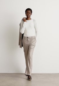 White long-sleeve top, beige plaid trousers with front pockets, and dark shoes. Plain background and subtle texture in the fabric.