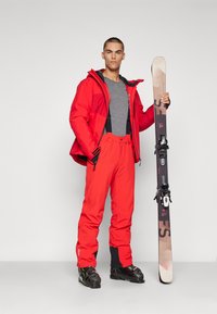 Chaqueta y pantalones de esquí rojos, camiseta gris; sosteniendo esquís con fijaciones. La chaqueta tiene cremalleras y capucha, y está hecha de un tejido texturizado; los pantalones tienen bolsillos.