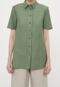 Camicia verde a maniche corte con chiusura a bottoni, caratterizzata da cuciture marrone chiaro lungo le cuciture e un colletto strutturato.