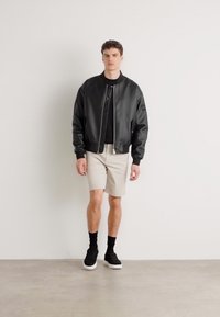 Black leather bomber jacket, short beige shorts, black socks, and black sneakers. Model stands against a plain white background.