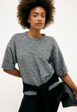 Junge Frau mit natürlichem lockigem Haar trägt einen grauen kurzärmeligen Strickpullover und hält eine schwarze gefaltete Jacke oder Tasche an ihrer Seite.