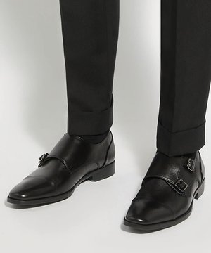 Black leather double monk strap dress shoes worn with black tailored pants against a light background.