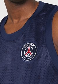 Sportovní tielko v námořnické modři s texturovanou síťovinou. Má kulatý výstřih a kulaté logo s nápisem "PARIS" a "SAINT-GERMAIN".