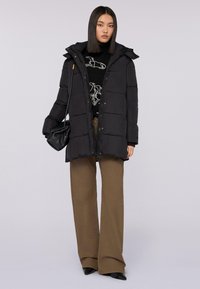 Schwarze Daunenjacke mit Reißverschluss und Kapuze, mit gesteppten Bereichen. Das Model trägt dunkle Hosen und hält eine kleine schwarze Handtasche.