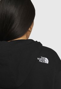 Svart hoodie med en åtsittande design, texturerat tyg och en vit broderad logotyp av "The North Face" på vänster axel.