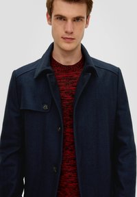 Marineblauwe wolmix jas met een visgraatpatroon, voorzien van een spreidkraag en drie knopen, gelaagd over een gestructureerde rode sweater.