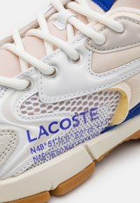 Lacoste sneakers feature a combination of white, beige, and blue materials, with mesh accents, thick white laces, and printed details in purple.