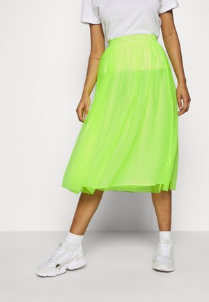 Personne portant une jupe midi vert néon vif, un t-shirt blanc, des chaussettes blanches et des baskets blanches, debout les jambes écartées sur un fond uni.