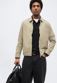 Άνδρας με μπεζ μπουφάν πάνω από μαύρο πουκάμισο και μαύρο τζιν, κρατώντας μαύρη δερμάτινη τσάντα στο δεξί χέρι μπροστά σε απλό φόντο.