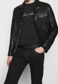 Black leather jacket with two chest pockets, layered over a dark gray sweatshirt with white text. Paired with dark denim pants and a black belt.