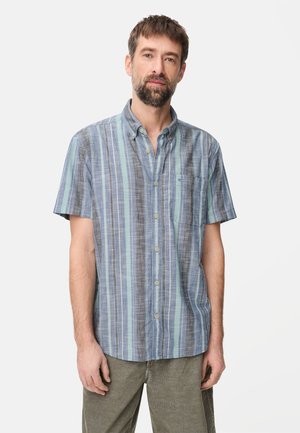 Mann mit Bart und kurzem Haar, der ein kurzärmeliges Hemd mit blauen, grauen und grünen Streifen sowie olivgrüne Hosen trägt.