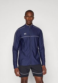 Μπλε ναυτικό αθλητικό μπουφάν με όρθιο γιακά, πλήρη φερμουάρ και λευκές λεπτομέρειες περιγράμματος. Το ύφασμα φαίνεται λείο με εφαρμοστή σχεδίαση. Λογότυπο μάρκας στο στήθος.