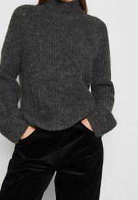 Pull en maille gris à col roulé avec texture côtelée et manches longues, associé à un pantalon en velours côtelé noir avec des rayures verticales et des poches.