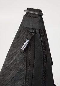 Geantă sling neagră cu o suprafață texturată, compartimente cu fermoar și un eticheta cu logo-ul Puma. Include un model geometric și o curea durabilă.