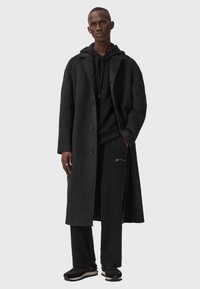 Schwarzer Wollmantel mit einreihigem Schnitt, kombiniert mit einem schwarzen Hoodie und passenden Hosen, getragen mit schwarzen Turnschuhen.