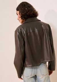Veste en cuir marron au design court, avec de larges revers et une texture lisse. Associée à un jean bleu clair, montrant la vue de dos et un style décontracté.