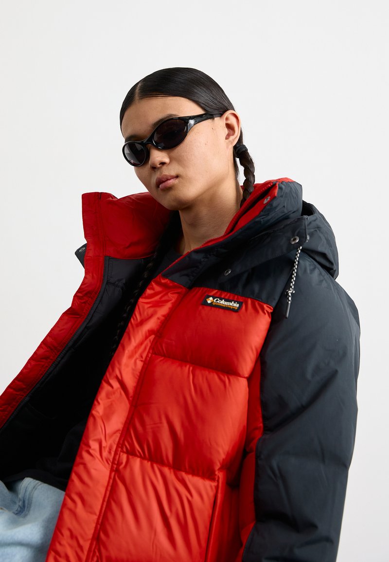 Rote und schwarze Steppjacke mit Kapuze, ausgestattet mit einem gesteppten Design und dem Columbia-Logo. Getragen mit schwarzen Sonnenbrillen.