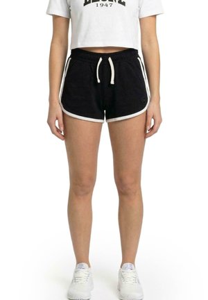 Modèle portant un short de sport noir à bordure blanche et à cordon de serrage, un T-shirt blanc court et des baskets blanches sur un fond uni.