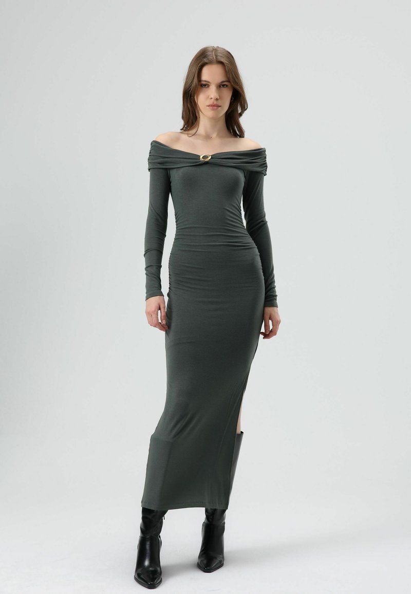 Robe à épaules dénudées à manches longues, en jersey vert foncé, avec un accent doré au niveau de l'encolure et des fentes latérales, portée avec des bottes noires.