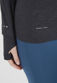 Långärmad, mörkgrå topp med texten "QUICK DRY" och hexagonala detaljer. Matchad med blå leggings med kontrasterande sömdetaljer.