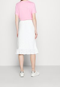 Weißer Stoffrock mit Lochmuster und Rüschensaum, kombiniert mit einem rosa T-Shirt mit kurzen Ärmeln und weißen Sneakers mit silbernen Akzenten.