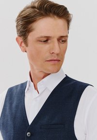 Granatowa kamizelka z teksturowanym splotem, zapięcie na jeden guzik i kieszeń, noszona na białej koszuli, prezentująca dopasowany krój.