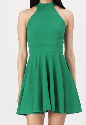 Kobieta ubrana w zieloną, bezrękawną sukienkę z wysokim kołnierzem, dopasowanym górnym elementem i rozkloszowanym dołem na jednolitym tle.