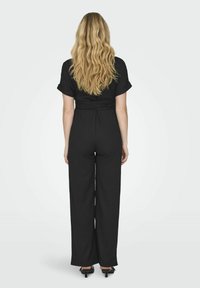 Mono negro de pierna ancha con mangas cortas y detalle de cinturón en la cintura. Textura de tela suave. Modelo de espaldas con el cabello ondulado.