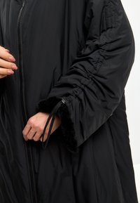 Sort isoleret jakke med lynlås foran, rynkede ærmedetaljer og faux pelstrimler. Materialet ser glat og blødt ud.