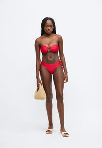 Piros bikinifelső, vállpánt nélküli, középen díszes karikával. Magas derekú bikini alsó, hozzá fonott kézitáska és semleges színű szandálok.