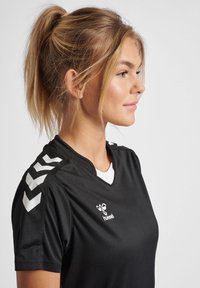 Black sports jersey with short sleeves, v-neck, white logo, and white chevron accents on shoulders. Smooth fabric texture.