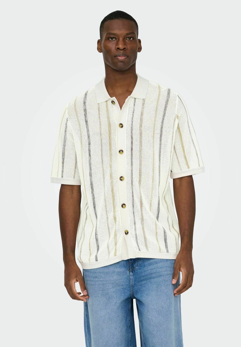 Homme portant une chemise blanche à manches courtes boutonnée avec des rayures verticales et un jean bleu clair, debout devant un fond uni.