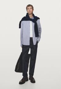Blue and white striped button-up shirt, white t-shirt, dark jeans, navy sweater draped, brown shoes, and a black bag. Model standing against white background.