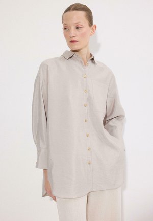 Femme portant une chemise beige clair oversize à boutons avec poche, manches longues, et pantalon couleur crème, debout devant un fond blanc.