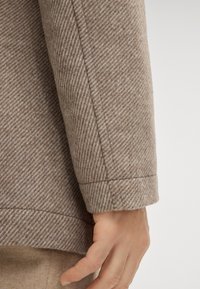 Rękaw beżowej wełnianej kurtki z wzorem jodełki, ukazujący czystą linię brzegu i subtelną fakturę, trzymany przez rękę o naturalnym odcieniu skóry.