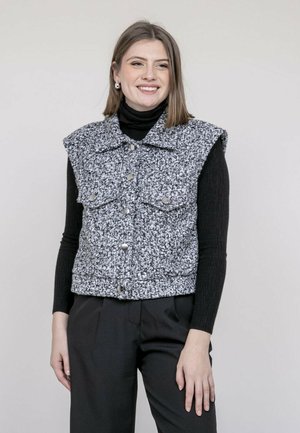 Femme portant un col roulé noir et un gilet sans manches texturé gris à boutons, debout devant un fond clair uni, souriante.