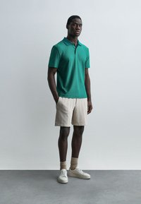 Άνδρας όρθιος με τα χέρια στις τσέπες, φορώντας πράσινη πουλόβερ πόλο με υφή, μπεζ σορτς, μπεζ κάλτσες και λευκά αθλητικά παπούτσια, πάνω σε απλό φόντο.