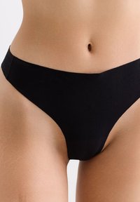 Torse inférieur d'une personne portant des sous-vêtements noirs sans coutures et taille haute sur fond clair.