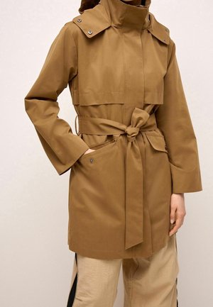 Trench-coat marron ceinturé avec poche à rabat à boutons-pression et capuche, porté sur un pantalon beige avec des bandes latérales noires.