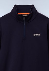 Sudadera de cremallera navy hecha de tejido suave, con un cuello alto, cremallera corta y un logo blanco en el pecho. Acento naranja brillante en el interior del cuello.