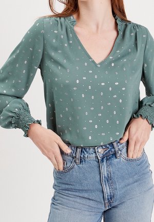 Blouse verte à manches longues avec motifs diamantés argentés, poignets smockés, col en V, rentrée dans un jean bleu clair taille haute avec les mains dans les poches.