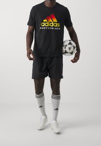 Osoba ubrana w czarną koszulkę Adidas Deutschland, czarne szorty, białe skarpety Adidas i białe korki trzyma czarno-białą piłkę nożną Adidas.