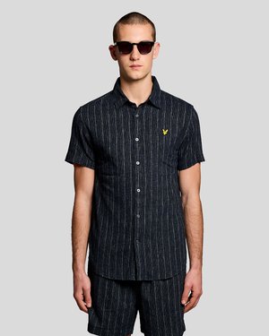 Mand iført mørk, stribet kortærmet skjorte med knapper og matchende shorts, accessoriseret med mørke solbriller på grå baggrund.
