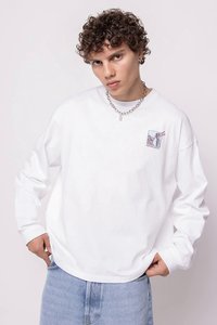 Jovem com cabelo encaracolado, vestindo uma camisa branca de manga longa com um pequeno gráfico e jeans azul claro, posando com as mãos nos bolsos.