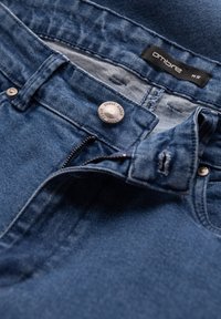 Vaqueros de mezclilla azul con una cremallera parcialmente abierta, botón de metal grabado con "OMBRE DENIM" y una etiqueta de marca "ombre" negra dentro de la cinturilla.
