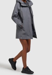 Osoba oblečená v šedé kapucínové deštníkce přes tmavé šaty, v černých sportovních botách, s rukama v kapsách bundy, kráčí proti neutrálnímu pozadí.