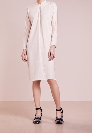 Kobieta w jasnobeżowej sukience do kolan z długim rękawem i stójką, zestawiona z czarno-białymi, teksturowanymi szpilkami na pasku wokół kostki.